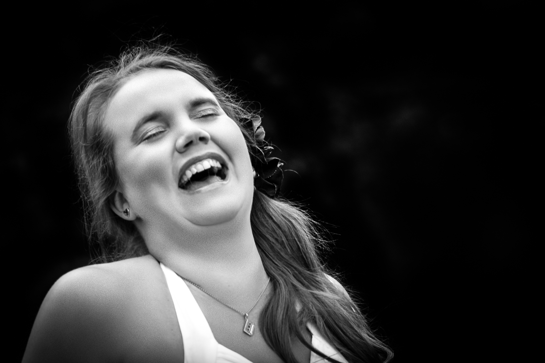 Black and white photo of a lady laughing with her eyes closed. She has a flower in her long hair over one shoulder, and is wearing a white halterneck dress. Her name is Dielle, pronounced Dee Ell.