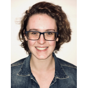 A head and shoulders picture of a smiling young woman in a jean jacket with short, curly brown hair, green eyes and glasses.